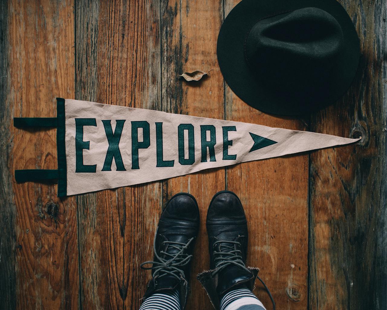 Top view of anonymous person wearing boots standing on wooden floor with Explore word on textile pointer placed near black hat